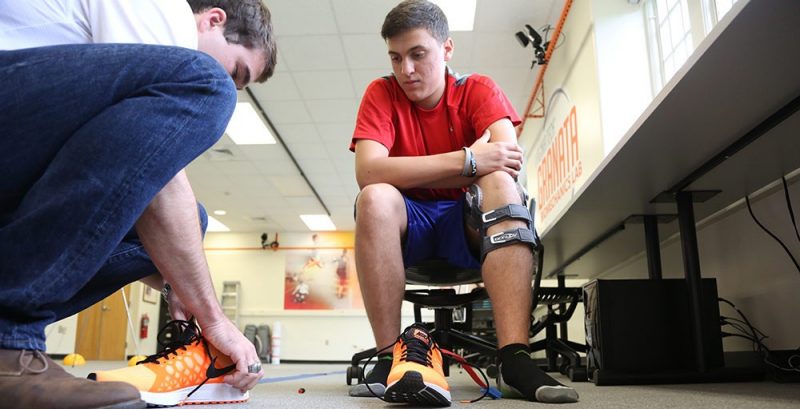 Boy sitting on a chair in the GRANATA laboratory with his shoes off while a lab member is placing pressure insoles into shoes